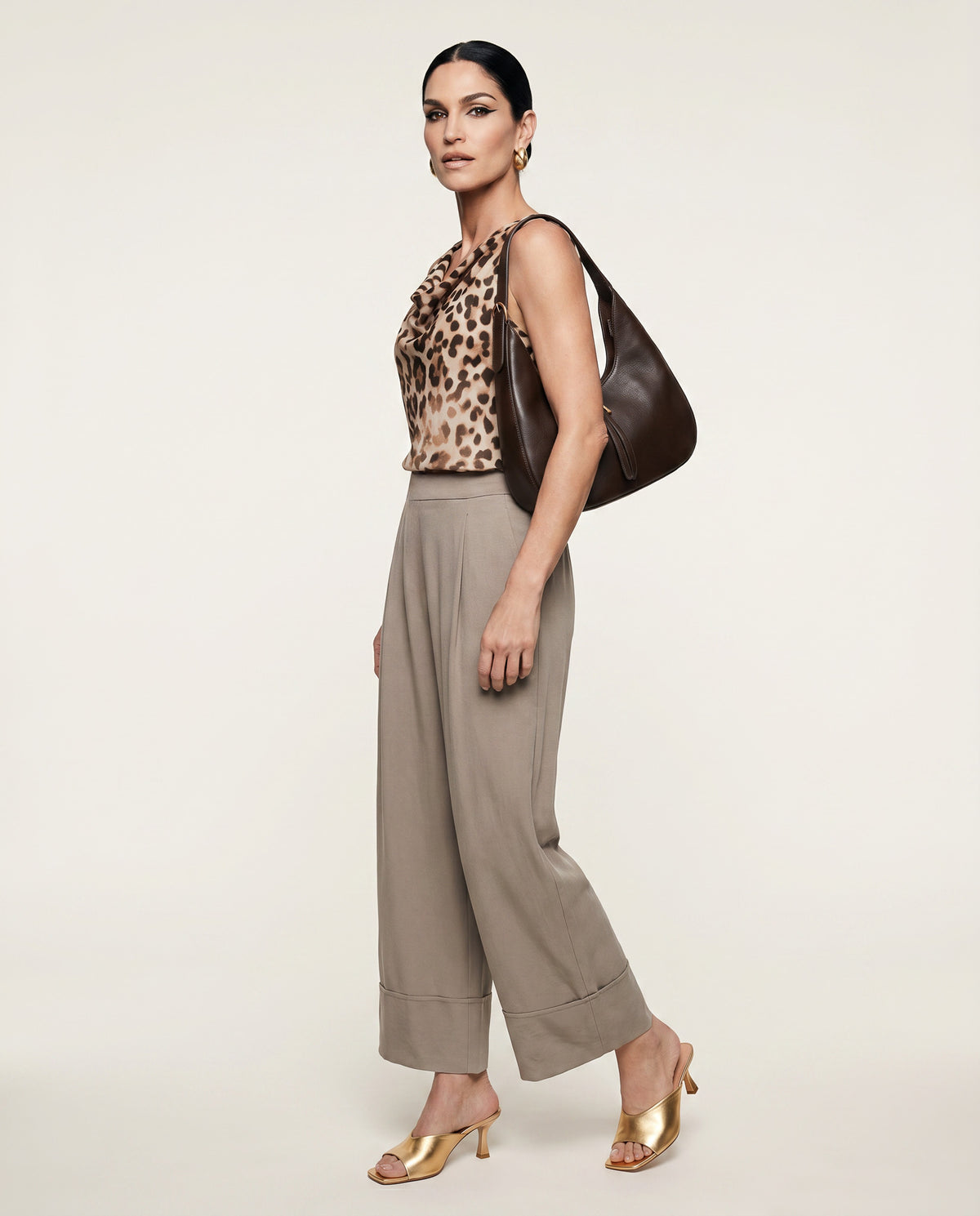 Woman wearing a leopard print top and beige pants, holding a brown handbag on a white background